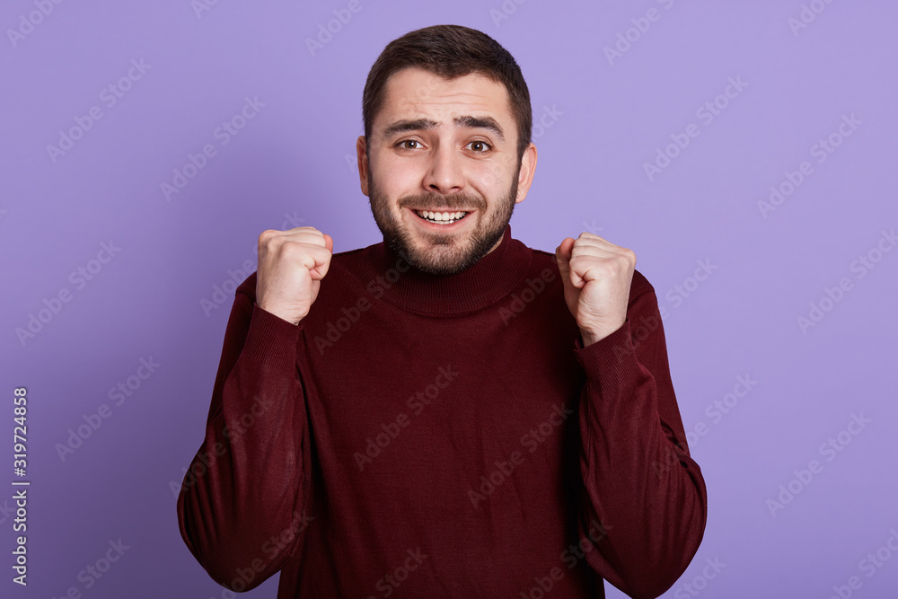 Fototapeta premium Indoor portrait of emotional charismatic good looking man with beard clenching fists, making gesture, smiling sincerely, having fun, getting good news, being in high spirits. People concept.