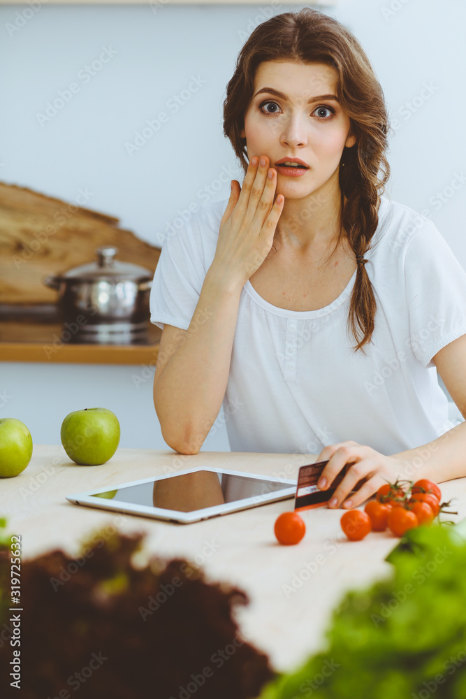Young woman looking for a new recipe for cooking in a kitchen. Housewife is making online shopping by tablet computer and credit card