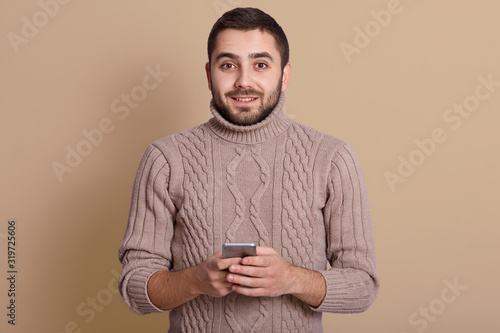 Image of pleasant positive young man with beard and black hair smiling sincerely, holding smartphone in both hands, looking directly at camera, having peaceful facial expression. Emotions concept.