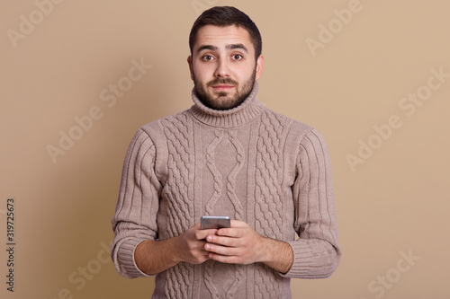 Picture of handsome magnetic man standing isolated over beige background, looking directly at camera, wearing beige warm sweater, holding smartphone in both hands. People and technology concept.