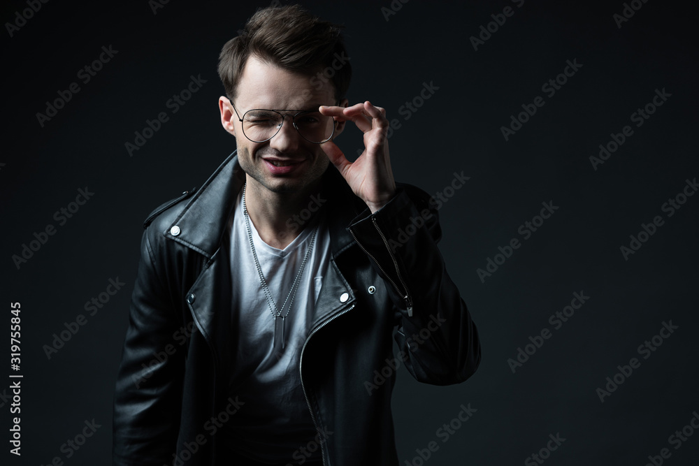 Obraz premium smiling stylish man in biker jacket touching eyeglasses and looking at camera isolated on black