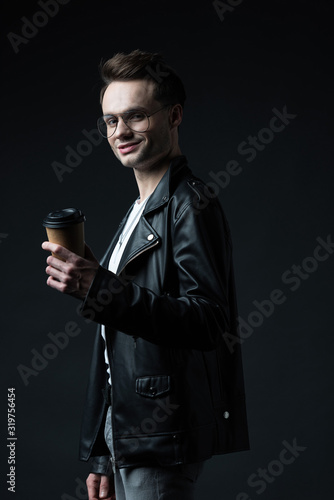 side view of smiling stylish brutal man in biker jacket with coffee to go isolated on black