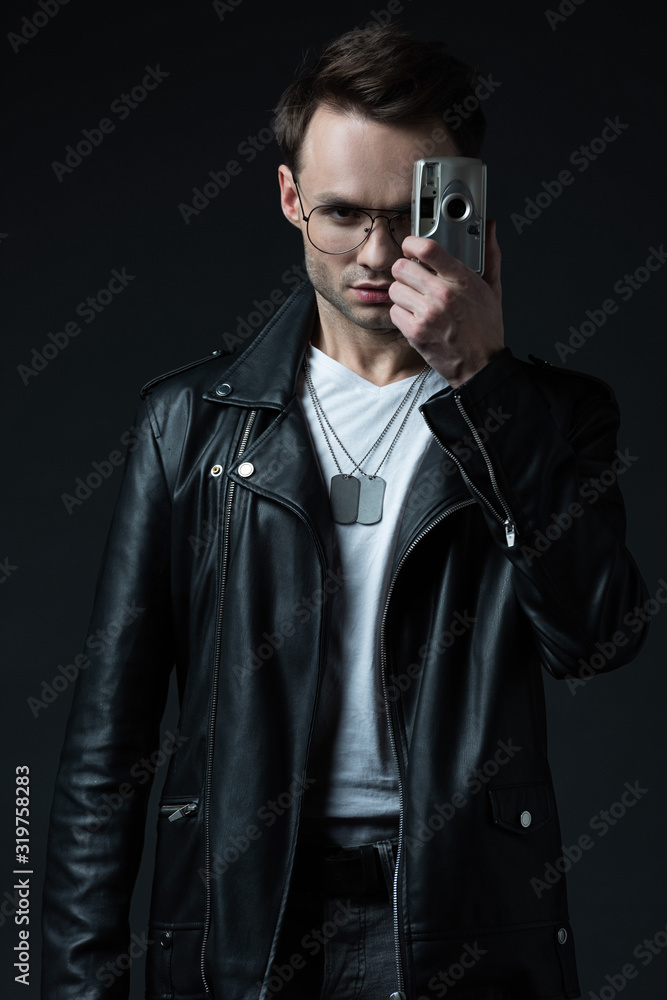 Obraz premium serious stylish brutal man in biker jacket taking picture on film camera isolated on black