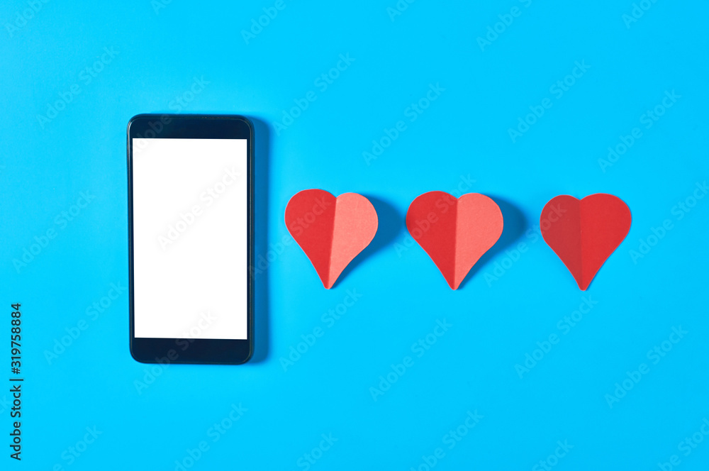 Black smartphone with isolated white screen for text, picture, photo and other graphics near row of three red paper hearts on blue table. Valentines day and love concept. Space for text. Top view Black smartphone with isolated white screen for text, picture, photo and other graphics near row of three red paper hearts on blue table. Valentines day and love concept. Space for text. Top view