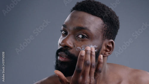 Portrait of attractive african young man using a moisturizing facial cream gently touching his face looking in the mirror. Close-up of nice-looking guy demostrating a jar of cream smiling at camera. © Fractal Pictures