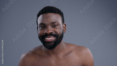 Close-up of attractive black topless gentleman looking at camera. Portrait of young man touching his black beard before shaving in the bathroom.