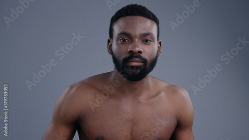 Naked closeup portrait of confident handsome african half naked man applying perfume on body isolated on grey background.