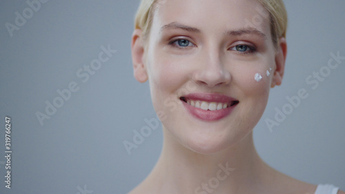 Close-up of stunning blonde young woman with perfect skin putting on moisturizing cream and smiling on camera against grey background. Skin care and cosmetics concept.