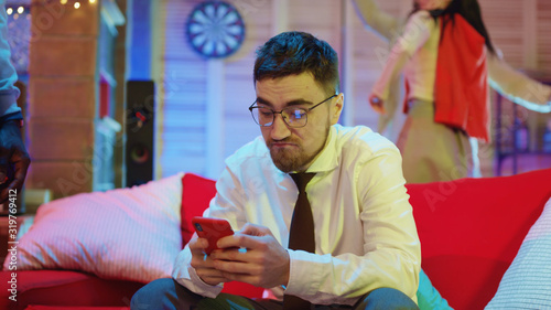 Handsome unsociable guy using social media or browsing news on smartphone getting upset on night party. People in background having fun celebration.