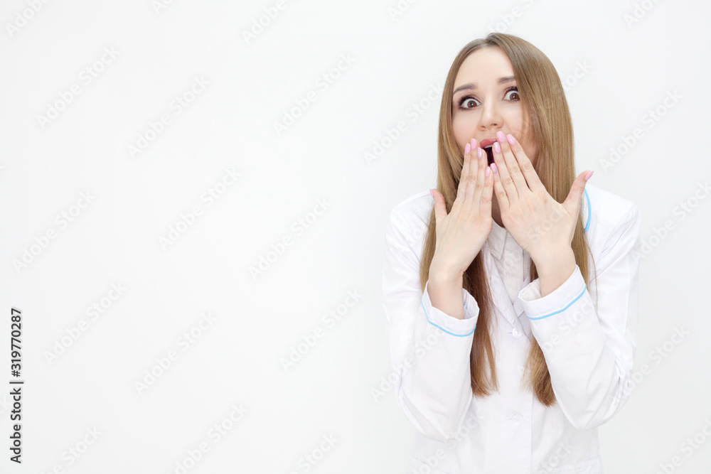 A girl in a medical uniform with an emotion of surprise, with wide eyes and mouth, covering the rum with her hand, on a white background. Concept of cosmetology and advertising of clinics. A girl in a medical uniform with an emotion of surprise, with wide eyes and mouth, covering the rum with her hand, on a white background. Concept of cosmetology and advertising of clinics.