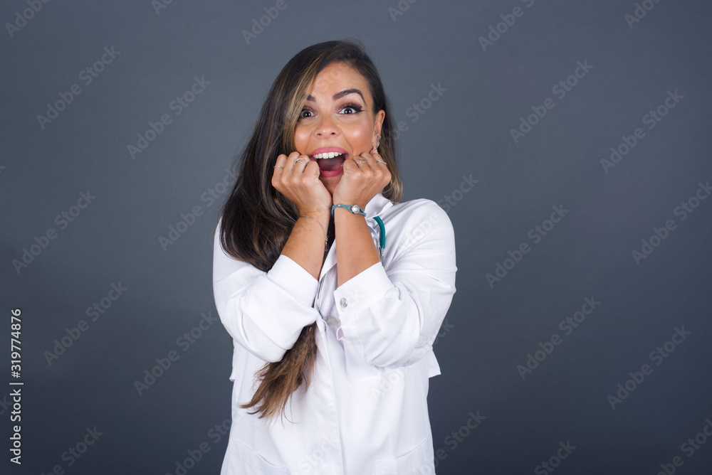 Studio shot of pleased lovely doctor woman keeps hands together under chin, being in high spirit, likes friends idea for next day, isolated on gray wall. Positive emotions. Studio shot of pleased lovely doctor woman keeps hands together under chin, being in high spirit, likes friends idea for next day, isolated on gray wall. Positive emotions.