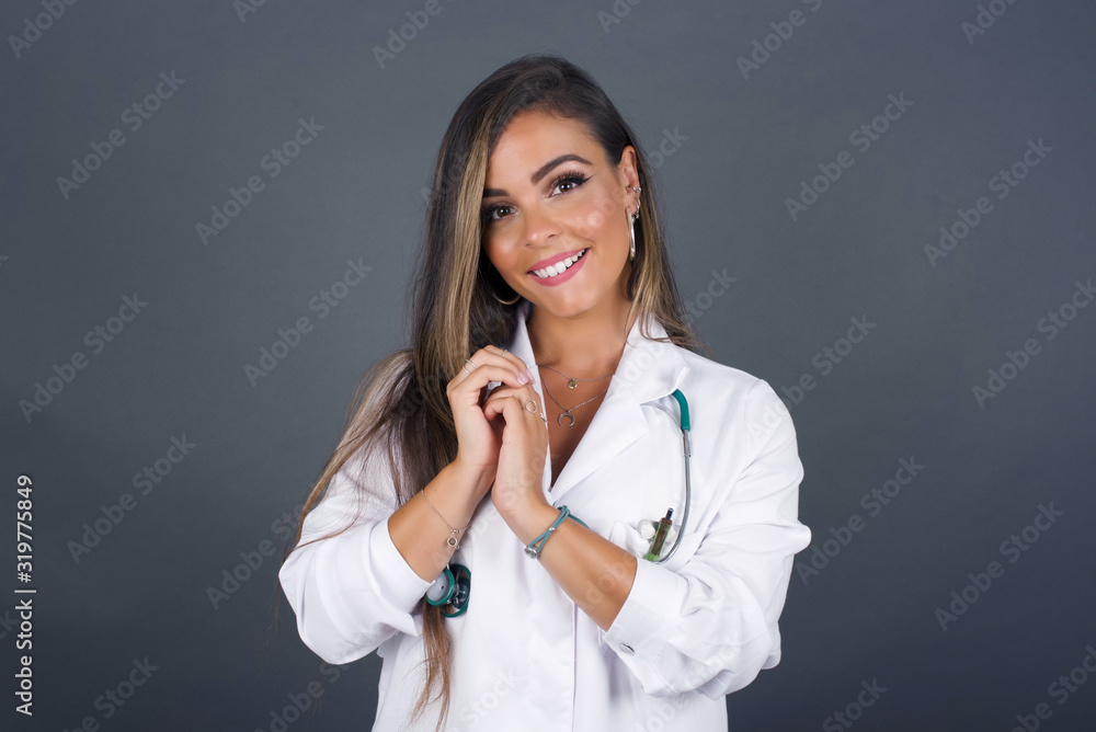 Dreamy charming young doctor female with pleasant expression, keeps hands crossed near face, excited about something pleasant, poses against gray background. Dreamy charming young doctor female with pleasant expression, keeps hands crossed near face, excited about something pleasant, poses against gray background.