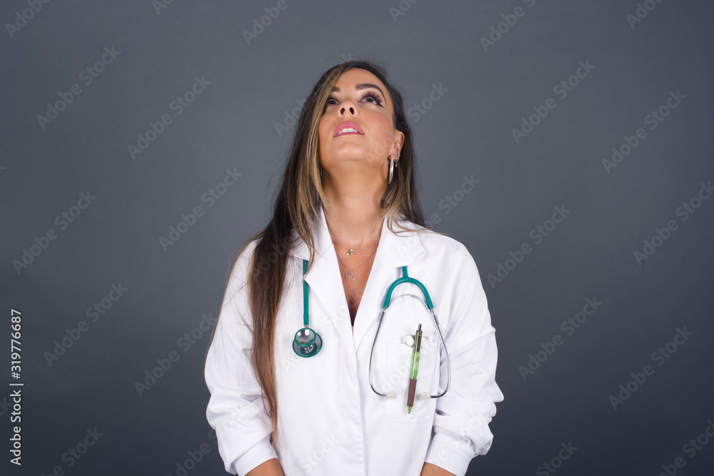 portrait of mysterious charming doctor woman with straight hair looking up with enigmatic smile. Beautiful smiling girl looking up standing against gray wall. portrait of mysterious charming doctor woman with straight hair looking up with enigmatic smile. Beautiful smiling girl looking up standing against gray wall.