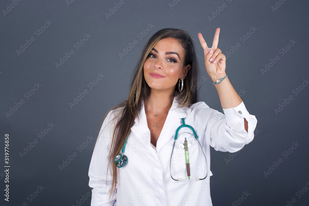 Young doctor woman wearing medical uniform standing against gray wall showing and pointing up with fingers number two or V-Sign while smiling confident and happy. Young doctor woman wearing medical uniform standing against gray wall showing and pointing up with fingers number two or V-Sign while smiling confident and happy.