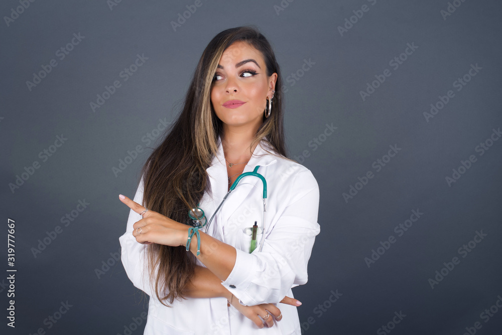 Pretty doctor woman posing on camera with tricky look and gesturing to side. Female designer presenting lovely product with index finger. Gesture concept. Pretty doctor woman posing on camera with tricky look and gesturing to side. Female designer presenting lovely product with index finger. Gesture concept.