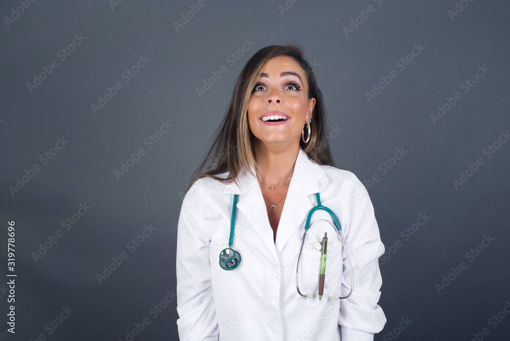 People and sudden reaction concept. Surprised caucasian beautiful doctor woman, shrugs shoulders, looking sideways, being happy and excited about new plans with friends. Isolated over gray wall. People and sudden reaction concept. Surprised caucasian beautiful doctor woman, shrugs shoulders, looking sideways, being happy and excited about new plans with friends. Isolated over gray wall.