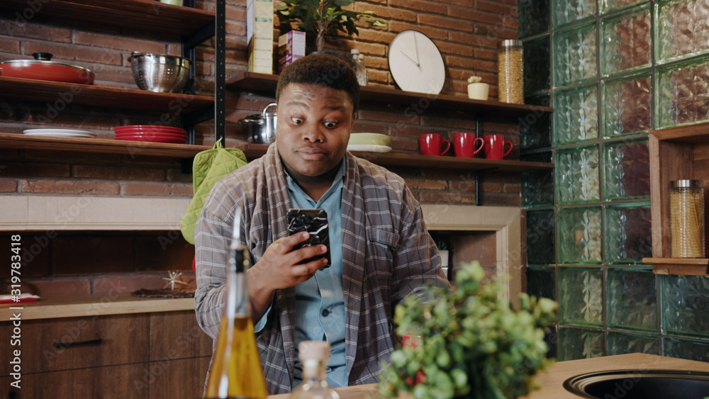 Handsome amazed black young man showing surprise face while looking on smartphone screen winning learning good news standing in the kitchen house interior.