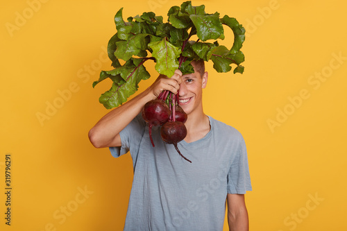 Horizontal shot of young handsome male holding bunch of beets in hand, hiding behind fresh vegetables, posing in casual outfit, covering hish face with beetroots. Raw food and healthy eating concept. photo