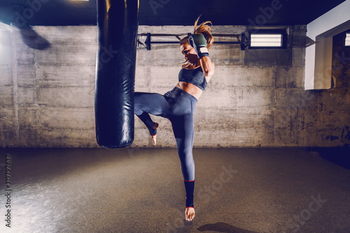 Young fit attractive muscular caucasian female kick boxer in sportswear with boxing gloves kicking boxing bag with knee.