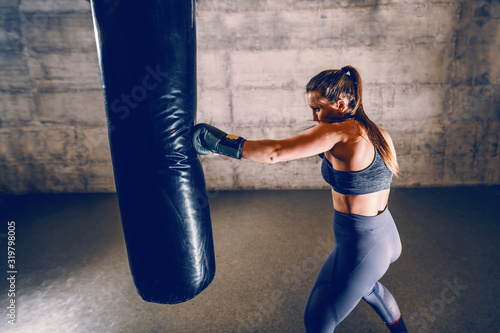 Young dedicated caucasian muscular female boxer in sportswear with ponytail and boxing gloves punching boxing bag while standing in the gym.