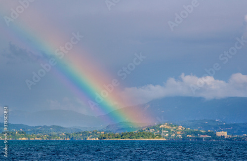 Beautiful summer landscape with sea  small village on the hill  bright colorful rainbow on blue sky  clouds and mountains on the horizon. Corfu Island  Greece