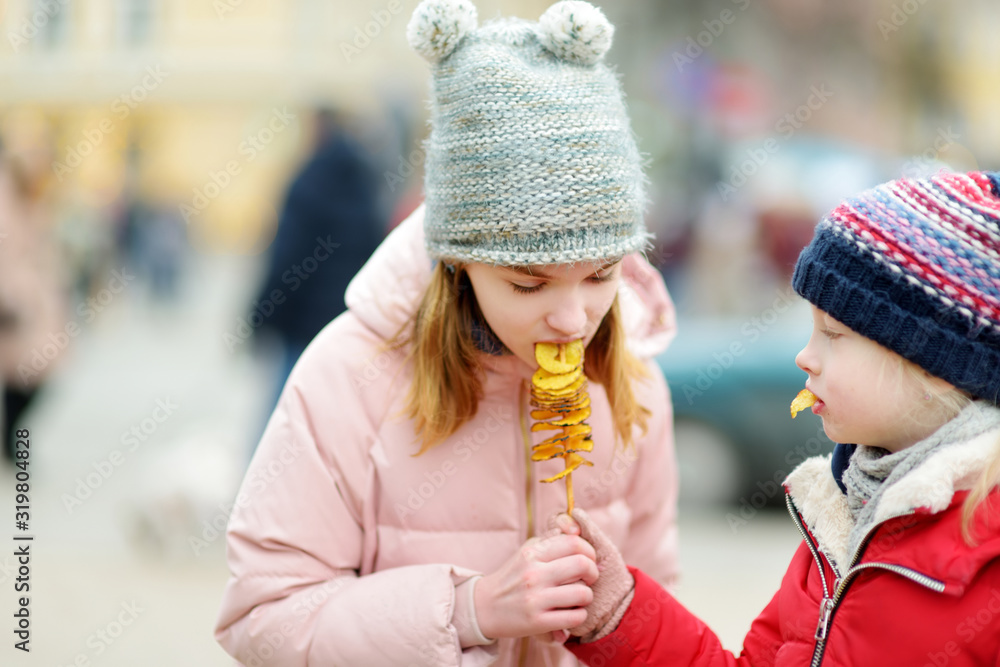 Cute little girls eating fried potato on a stick on cold winter day outdoors. Cute little girls eating fried potato on a stick on cold winter day outdoors.