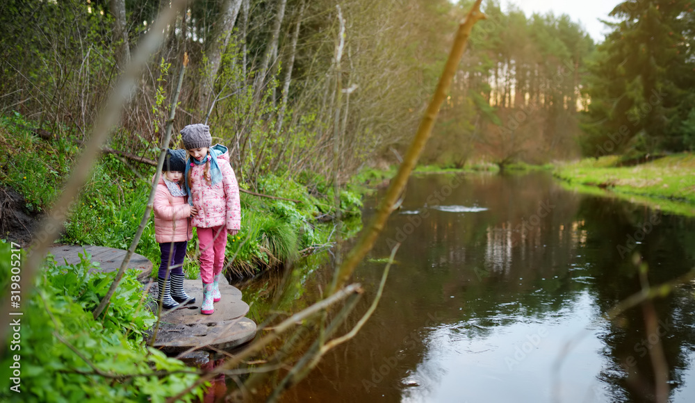 Two cute young sisters having fun by a river on warm spring day. Children playing together by a water. Two cute young sisters having fun by a river on warm spring day. Children playing together by a water.
