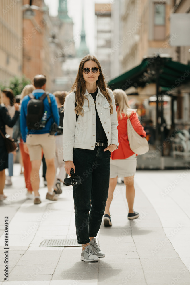 A gorgeous young female blogger in a white jeans jacket, black jeans, grey sneakers, and sunglasses holds the tripod with smartphone poses in the center of ancient European street full of people