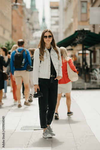 Obraz na płótnie A gorgeous young female blogger in a white jeans jacket, black jeans, grey sneakers, and sunglasses holds the tripod with smartphone poses in the center of ancient European street full of people w salonie