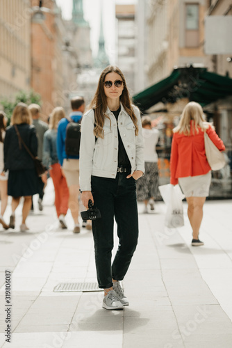 A gorgeous young female blogger in a white jeans jacket, black jeans, grey sneakers, and sunglasses holds the tripod with smartphone poses in the center of busy ancient European street