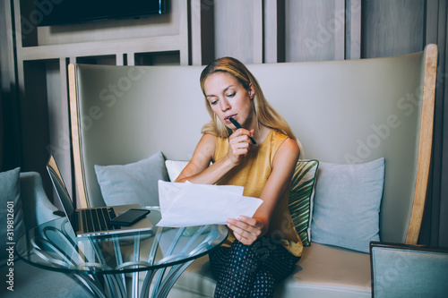 Fotografie Concentrated blonde woman in smart casual wear pondering while checking planning strategy for project holding paper, millennial female manager checking data using modern technologies for research