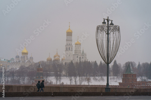 Winter city downtown. Photography of hurrying up pair of young man and woman on the street within snowfall. Kremlin Towers and Ivan the Great Bell Tower as background. 