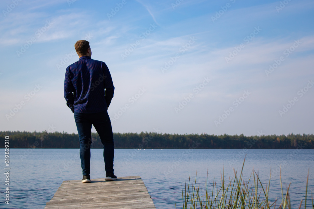 A man stands on a wooden bridge on a lake on a clear day. Blue sky and lake. Scenery A man stands on a wooden bridge on a lake on a clear day. Blue sky and lake. Scenery