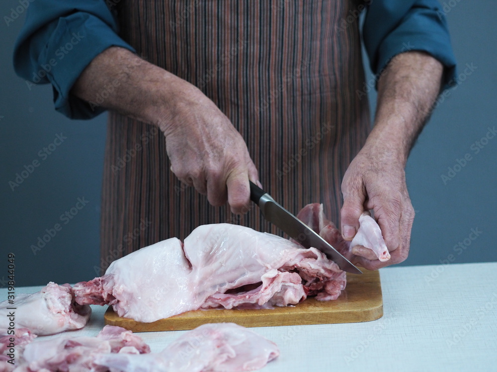 A male chef holds a knife and cuts into pieces raw rabbit meat on a cutting kitchen board.
