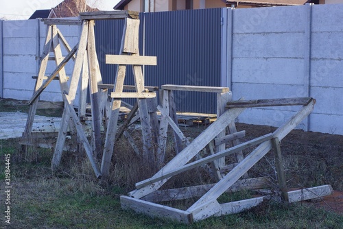 a pile of gray wooden construction goats stands on the green grass on the street at a concrete fence and an iron gate