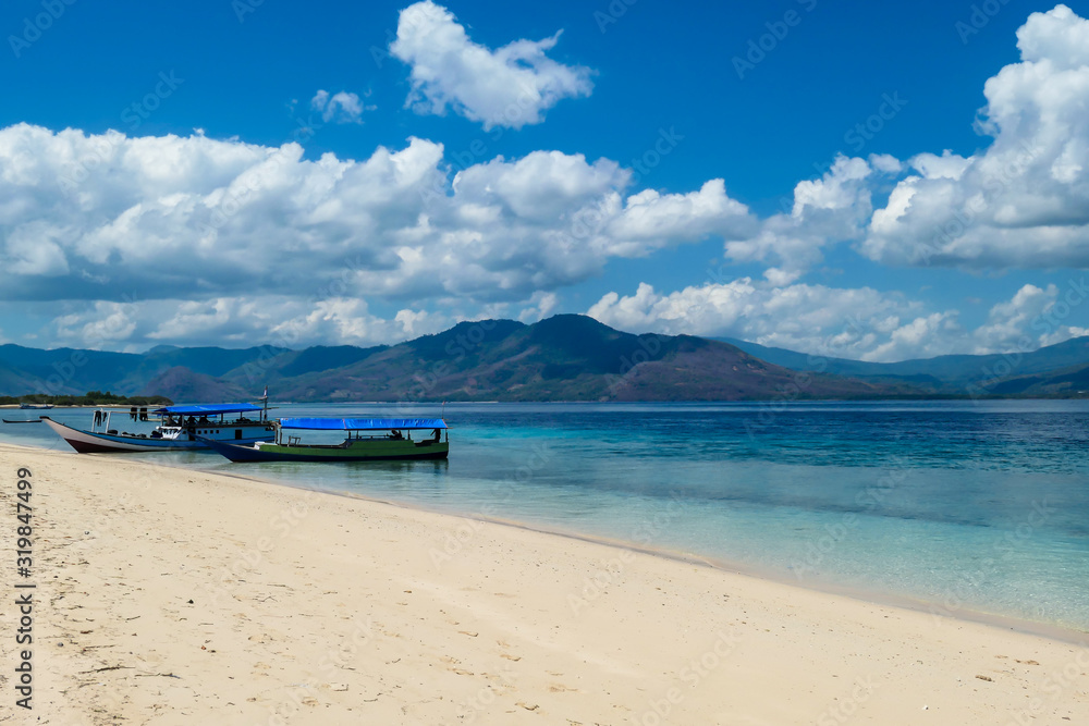 Two boats anchored to white sand beach on an island near Maumere, Indonesia. There is a fishing cottage on the side. Clear, turquoise coloured water displaying coral reef