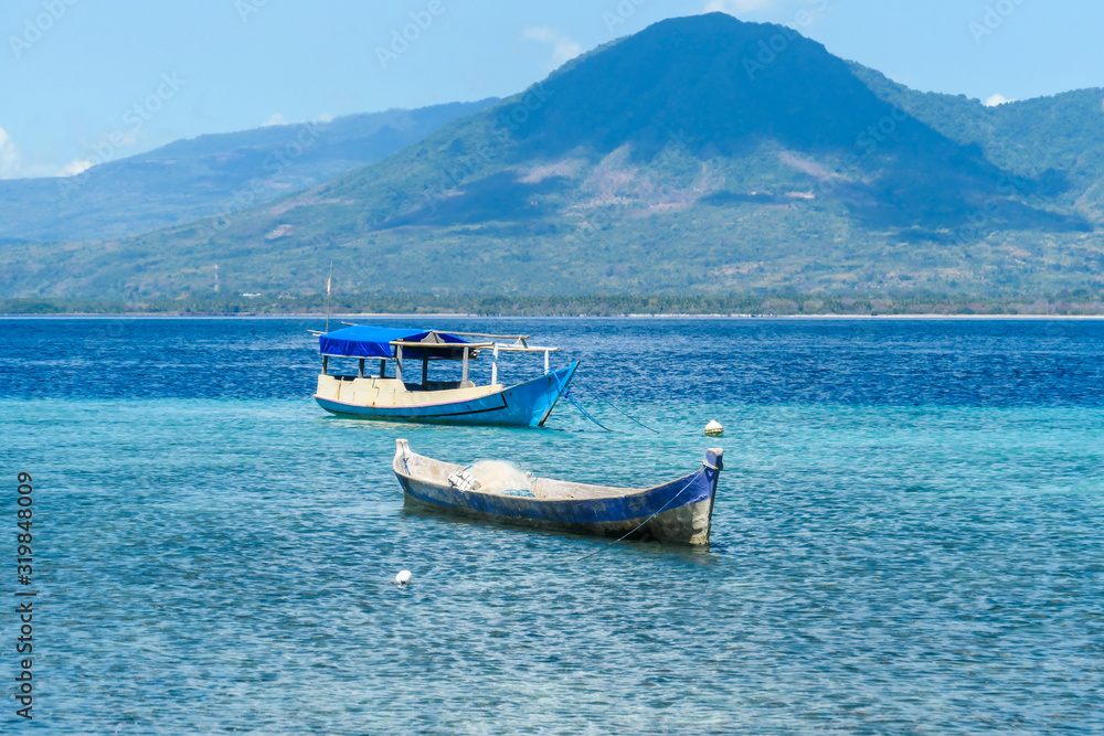 Two boats anchored to shallow waters of a small island near Maumere, Indonesia. Clear, turquoise coloured water displaying coral reef. There is a massive island in the back. Serenity and calmness.