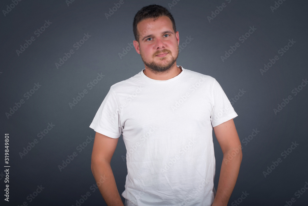 Confidence and business concept. Portrait of charming successful young man, smiling broadly with self-assured expression while holding hands in pockets standing against gray wall. Confidence and business concept. Portrait of charming successful young man, smiling broadly with self-assured expression while holding hands in pockets standing against gray wall.