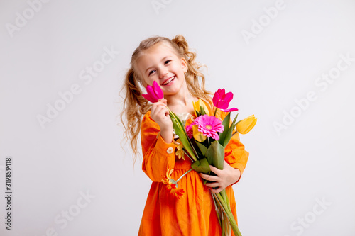 little blonde girl in an orange dress holds tulips and gerberas in her hands on a white background, a child girl smiles and holds spring flowers in her hands, space for text