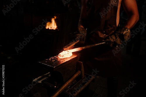 Brutal bearded peasant craftsman banging on red hot metal on an anvil with a hammer in a dark workshop