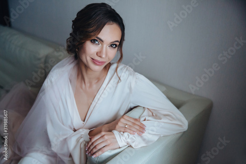 Morning bride, beautiful bride is waiting for the wedding sitting on the sofa in the white room. Girl in wedding dress in front of the window is waiting for the groom photo