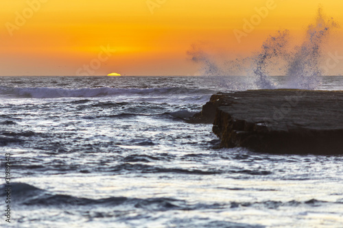 An amazing view of the sunset over the water in the Chilean coast. An idyllic beach scenery with the sunlight illuminating the rocks while the waves are splashing water drops to the sky 