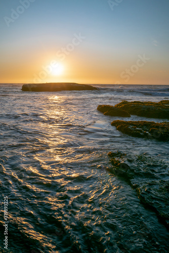 An amazing view of the sunset over the water in the Chilean coast. An idyllic beach scenery with the sunlight illuminating the green algae and rocks with orange tones and the sea in the background 