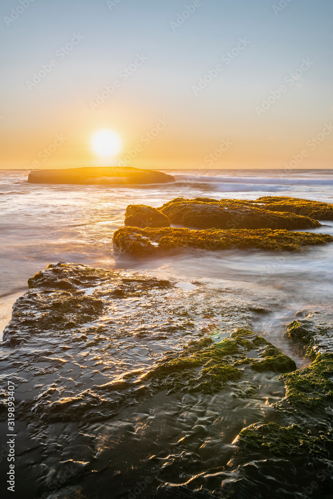 An amazing view of the sunset over the water in the Chilean coast. An idyllic beach scenery with the sunlight illuminating the green algae and rocks with orange tones and the sea in the background

