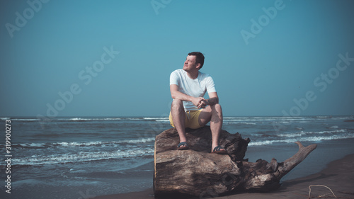 Relaxed man sits on log and looks away. Male tourist sits on driftwood and squints his eyes at bright sun  resting on sandy beach near waving sea.