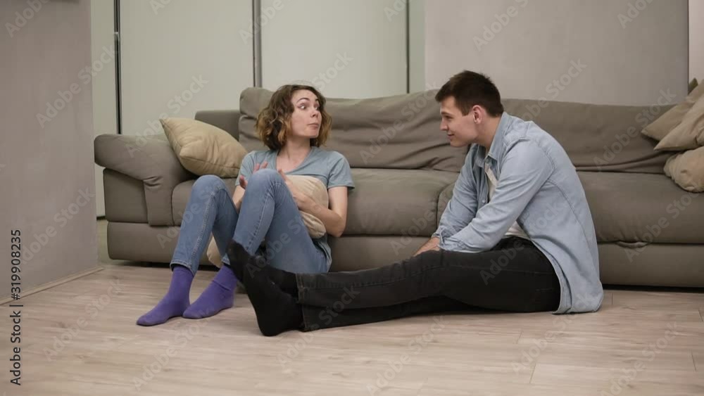Upset stressed young caucasian girl feeling offended by boyfriend or husband while he is smiling - arguing shouting, jealousy. Sitting on the floor. Small quarrel in marriage relationship Upset stressed young caucasian girl feeling offended by boyfriend or husband while he is smiling - arguing shouting, jealousy. Sitting on the floor. Small quarrel in marriage relationship