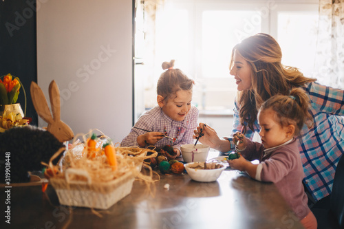 Two female toddlers and their mother painting eggs for easter in the kitchen.