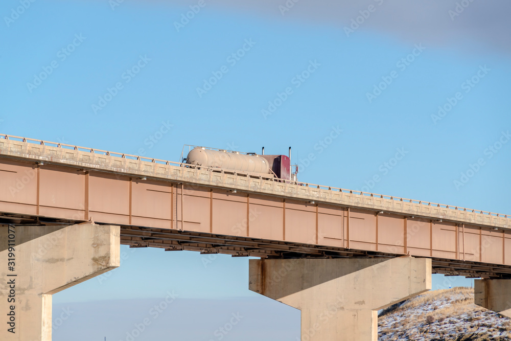 Truck with large tank container travelling on a bridge over lake on a sunny day
