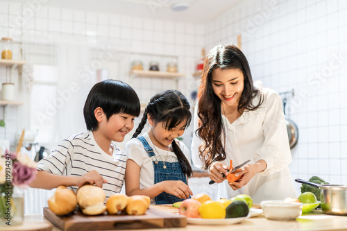 Lovely cute Asian family making food in kitchen at home. Portrait of smiling mother and children standing at cooking counter preparing ingredient for dinner meal. Happy family activity together.