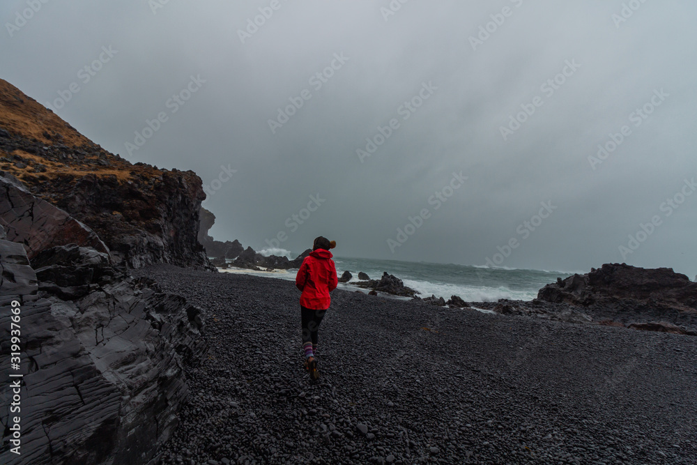 Obraz premium Girl with a red rain coat and a biene hat on Djupalonssandur black sand beach